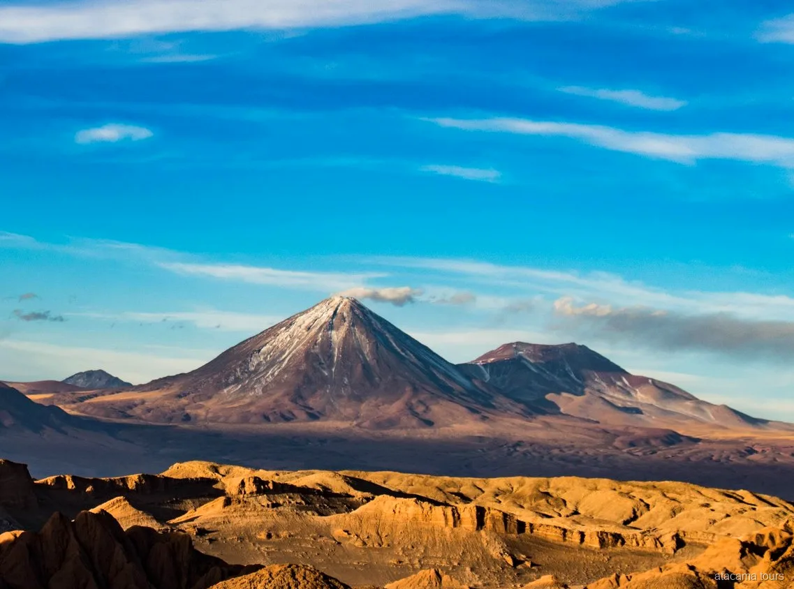 Paysage des excursions dans l'Atacama, cône volcanique enneigé dominant des crêtes désertiques dorées