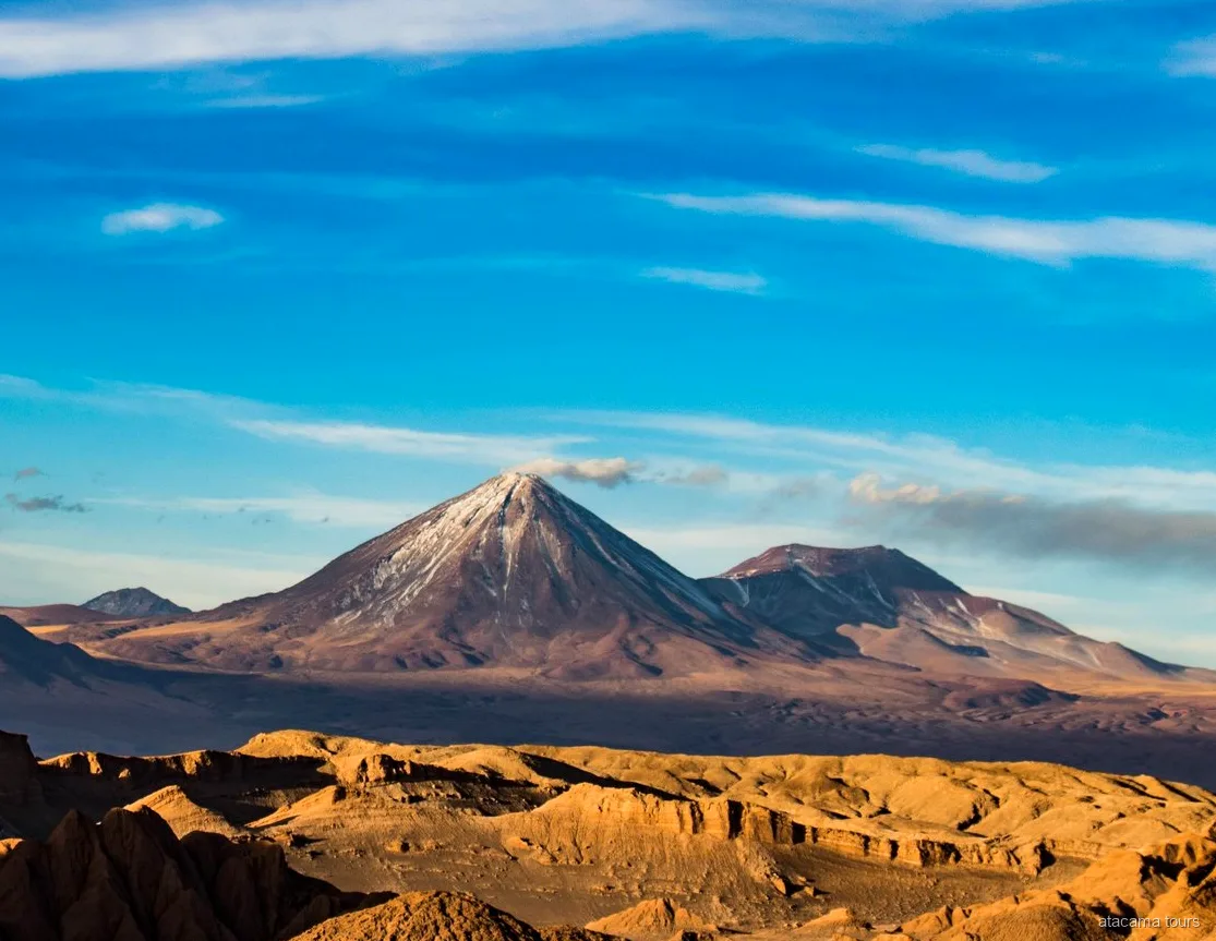 Landschap op Atacama tours, besneeuwde vulkaankegel boven goudkleurige, geërodeerde woestijnruggen