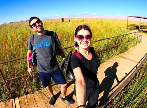 Stel op Atacama tours maakt een selfie op een houten vlonderpad door hoog gras, vulkanen aan de horizon