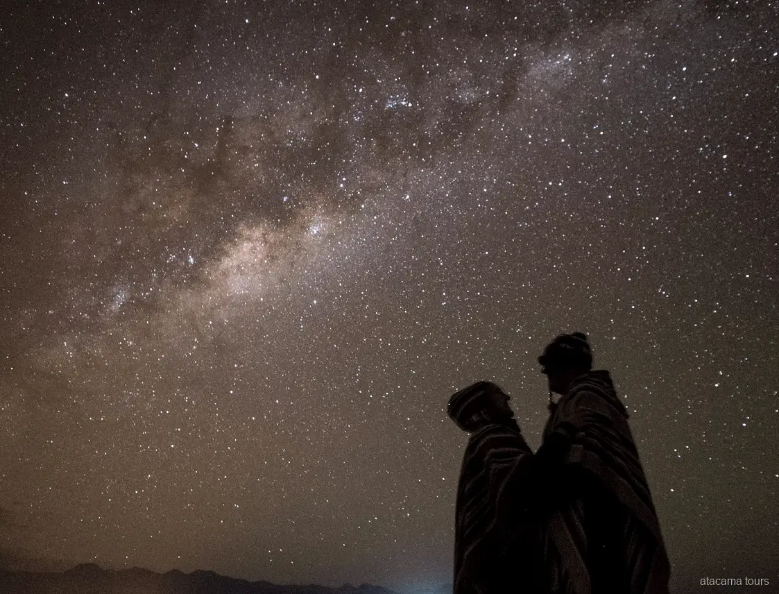 Tours astronómicos en Atacama, dos siluetas envueltas en ponchos bajo una densa franja de la Vía Láctea
