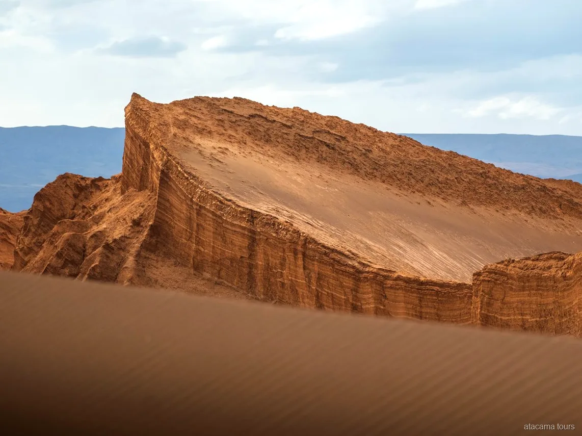 Tour nel deserto di Atacama al Valle de la Luna, parete rocciosa inclinata a strati sopra una duna di sabbia