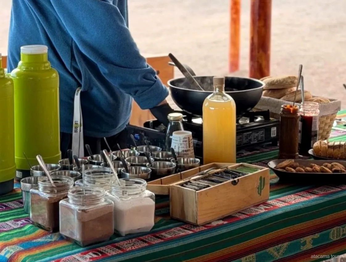 Mesa de refrigerios de los tours en Atacama: termos, frascos, pan y un hornillo sobre un mantel andino a rayas