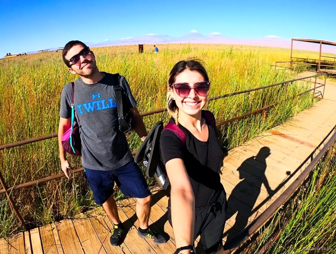 Coppia in tour nel deserto di Atacama, selfie su una passerella di legno tra l'erba alta, vulcani all'orizzonte